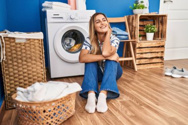 Young blonde woman sitting on the floor waiting for washing machine at laundry room