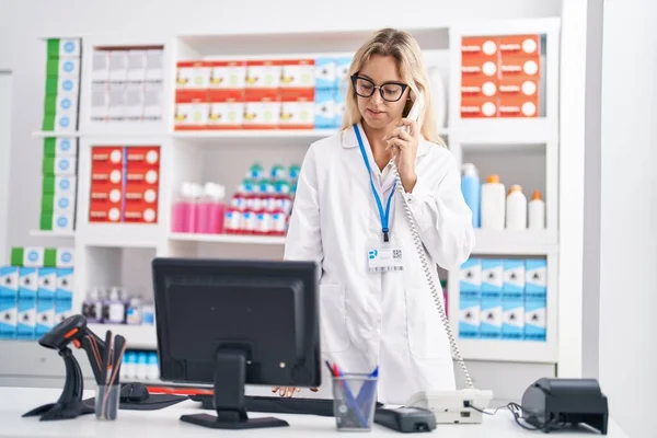 若いブロンドの女性の薬剤師は薬局でコンピュータを使って電話で話す — ストック写真