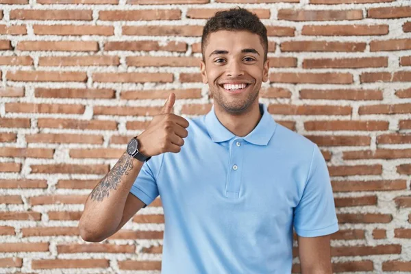 手でジェスチャーを幸せな親指を行うレンガの壁の上に立ってブラジルの若い男 成功を示すカメラを見ると — ストック写真