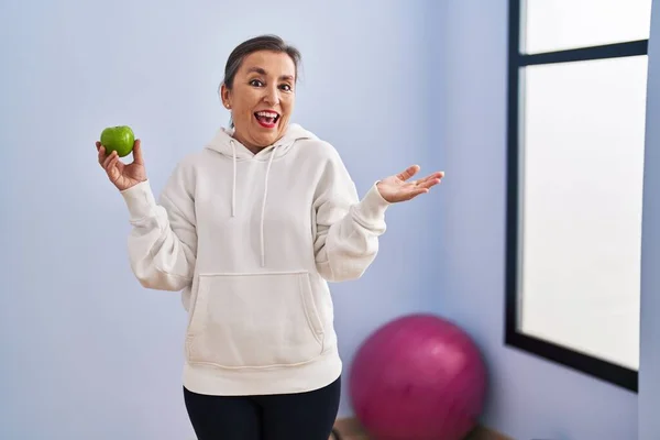 健康的なアップルを保持しているスポーツウェアを身に着けている中年のパニック女性幸せな笑顔と受賞式で手を上げ — ストック写真