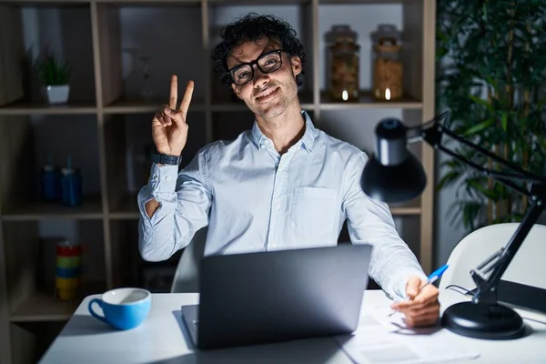 Hispanic man working at the office at night smiling looking to the camera showing fingers doing victory sign. number two.