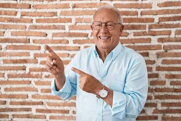 stock image Senior man with grey hair standing over bricks wall smiling and looking at the camera pointing with two hands and fingers to the side. 