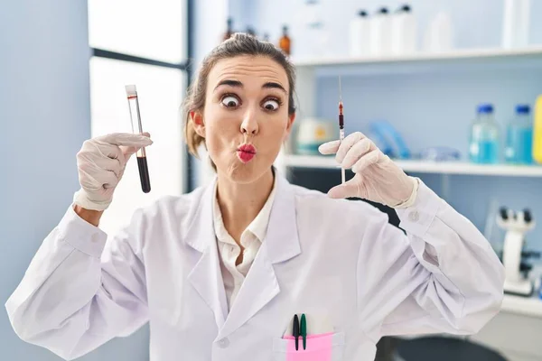 Young woman working at scientist laboratory holding blood sample making fish face with mouth and squinting eyes, crazy and comical.