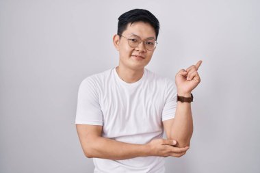 Beyaz arka planda duran genç Asyalı adam yüzünde büyük bir gülümsemeyle, kameraya el ile işaret ederek bakıyor.. 