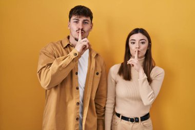 Sarı arka planda dikilen genç İspanyol çift parmakları dudaklarında sessiz olmak istiyorlar. sessizlik ve gizli konsept. 