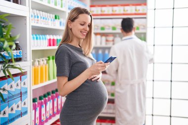 Erkek ve kadın eczacı ve hamile müşteri eczanede akıllı telefon kullanarak konuşuyor.