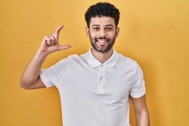 Sarı arka planda duran Arap adam gülümsüyor ve el hareketleriyle hareket ediyor parmaklarıyla işaret yapıyor ve kameraya bakıyor. ölçüm kavramı. 