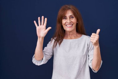 Orta yaşlı bir kadın mavi arka planda duruyor. Altıncı parmaklarını gösterip işaret ederken kendine güvenen ve mutlu bir şekilde gülümsüyor.. 