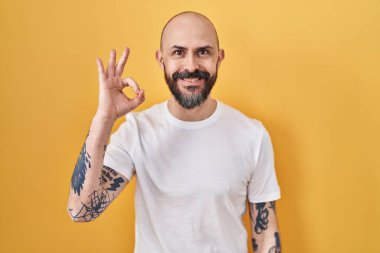 Sarı arka planda dövmeleri olan Latin asıllı genç adam gülümsüyor ve el ve parmaklarıyla 