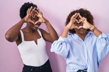 Pembe arka planda duran iki Afrikalı kadın. Elleri ve parmaklarıyla kalp şeklinde tabelanın içinden bakıp gülümsüyorlar. 