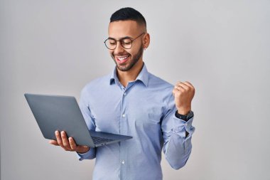 Genç İspanyol adam bilgisayarını kullanarak çok mutlu ve heyecanlı bir şekilde kollarını kaldırarak, gülümseyerek ve başarı için bağırarak kazanan jestler yapıyor. kutlama konsepti. 