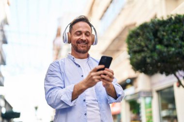 Genç, beyaz bir adam gülerek sokakta müzik dinliyor.