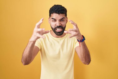 Hispanic man with beard standing over yellow background shouting frustrated with rage, hands trying to strangle, yelling mad 