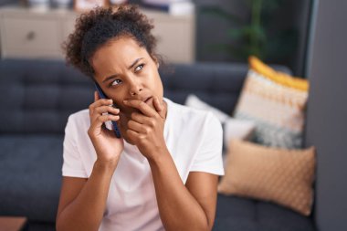 Afro-Amerikalı kadın evde endişeli bir ifadeyle akıllı telefondan konuşuyor.