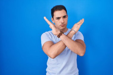 Young hispanic man standing over blue background rejection expression crossing arms doing negative sign, angry face 