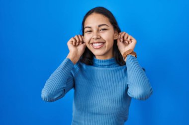 Mavi izole edilmiş arka planda dikilen genç Brezilyalı kadın parmaklarıyla kulaklarını çekerek gülümsüyor, komik bir jest. deneme sorunu 