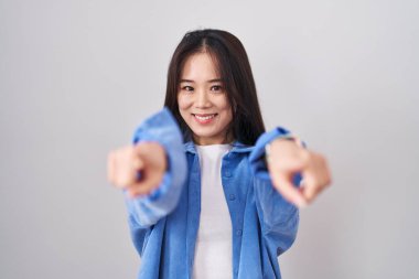 Beyaz arka planda duran genç Çinli kadın seni ve kamerayı işaret ediyor, pozitif ve neşeli gülümsüyor. 
