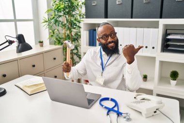 Tıbbi klinikte çalışan Afro-Amerikan bir adam elinde çekiçle baş parmağını kaldırıyor ve ağzı açık gülümsüyor. 