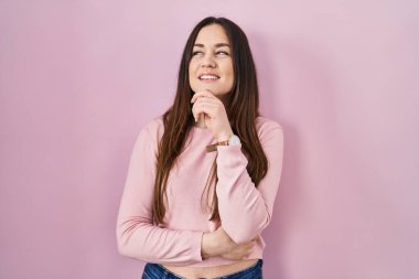 Young brunette woman standing over pink background with hand on chin thinking about question, pensive expression. smiling and thoughtful face. doubt concept. 