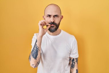 Sarı arka planda dikilen dövmeleri olan genç İspanyol bir adam ağzı ve dudakları parmaklarıyla kapanmış bir fermuar gibi. gizli ve sessiz, tabu konuşması 