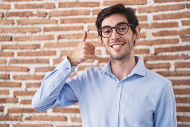 Duvarın arkasında dikilmiş gülümseyen İspanyol genç adam telefonla el ve parmaklarıyla telefon hareketi yapıyor telefonda konuşuyor gibi. iletişim kavramları. 