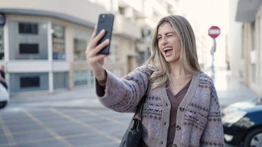 Young blonde woman smiling confident making selfie by the smartphone at street