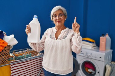 Beyaz saçlı, çamaşır ve deterjan şişesi yıkayan orta yaşlı bir kadın bir fikir veya soruyla gülümsüyor ve mutlu bir yüzle işaret ediyor. 