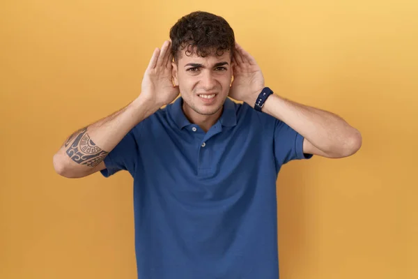 Молодой Латиноамериканец Стоящий Жёлтом Фоне Пытающийся Услышать Обе Руки Ушном — стоковое фото