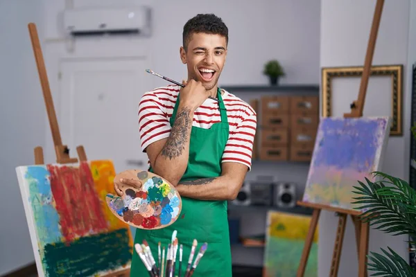 巴西年轻人拿着画家的调色板在艺术家工作室眨眼看着相机 带着性感的表情 开心而快乐的脸 — 图库照片