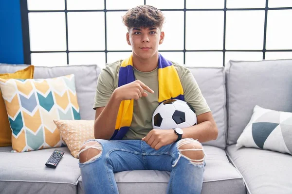 Hispanic teenager sitting on the sofa watching football match smiling happy pointing with hand and finger