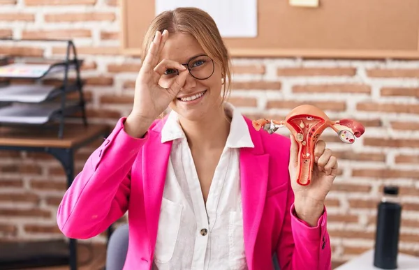 Young caucasian woman holding model of female genital organ at the office complaining for menstruation pain smiling happy doing ok sign with hand on eye looking through fingers