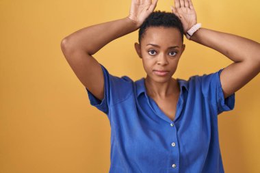 Afrika kökenli Amerikalı bir kadın sarı arka planda duruyor. Tavşan kulağı hareketi yapıyor. Avuçlarıyla alaycı ve şüpheci görünüyor. Paskalya tavşanı konsepti. 