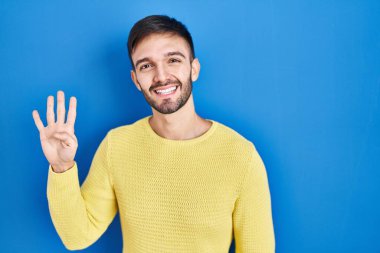 Hispanic man standing over blue background showing and pointing up with fingers number four while smiling confident and happy. 