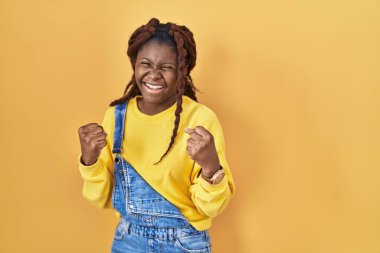 African woman standing over yellow background very happy and excited doing winner gesture with arms raised, smiling and screaming for success. celebration concept. 