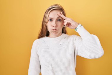 Sarı arka plan üzerine beyaz kazak giyen genç beyaz bir kadın. Alnındaki sorun yüzünden endişeli ve stresli. Kriz konusunda endişeli. 