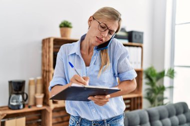 Young blonde woman psychologist writing on clipboard and talking on the smartphone at clinic