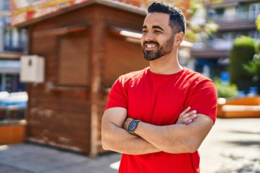 Genç İspanyol adam gülümsüyor. Parkta kollarını kavuşturmuş duruyor.