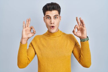 Mavi arka planda dikilen Latin asıllı genç adam şaşırmış ve şok olmuş görünüyor. Tamam sembolünü parmaklarıyla yapıyor. çılgın ifade 