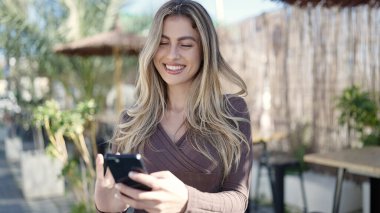 Young blonde woman smiling confident using smartphone at coffee shop terrace