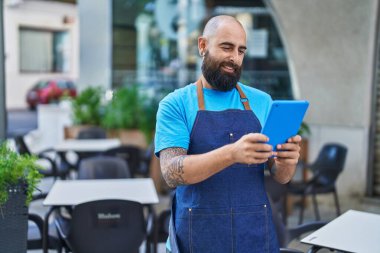 Young bald man waiter smiling confident using touchpad at coffee shop terrace