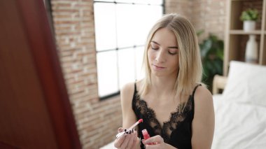 Young blonde woman putting lipstick sitting on bed looking sexy at the mirror at bedroom