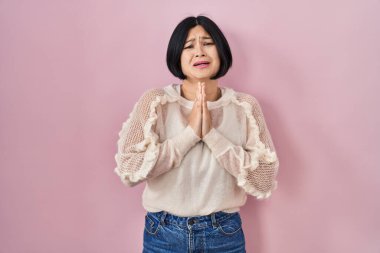 Young asian woman standing over pink background begging and praying with hands together with hope expression on face very emotional and worried. begging. 