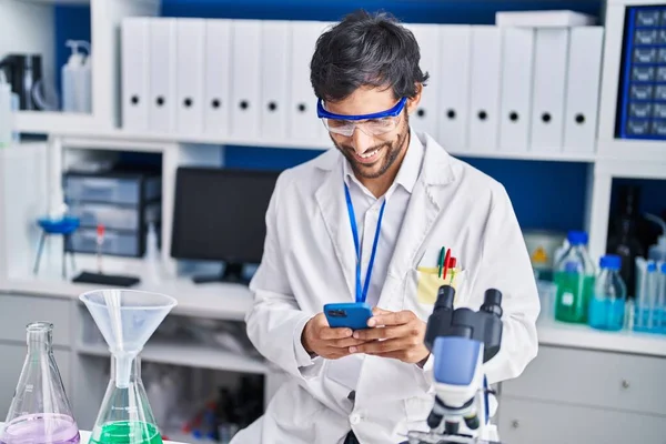 研究室でスマートフォンを使って自信を持って笑っている若いヒスパニック系の男性科学者 — ストック写真