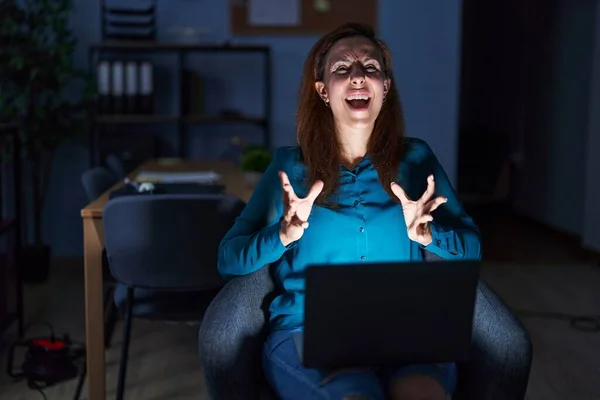 Brunette woman working at the office at night crazy and mad shouting and yelling with aggressive expression and arms raised. frustration concept.