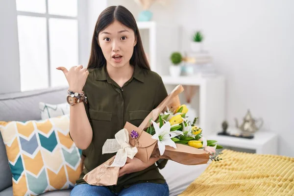 stock image Chinese young woman holding bouquet of white flowers surprised pointing with hand finger to the side, open mouth amazed expression. 