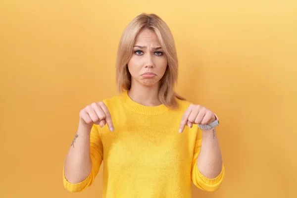 年轻的高加索女人穿着黄色毛衣 垂头丧气地垂头丧气 用手指指示方向 既不快乐又沮丧 — 图库照片