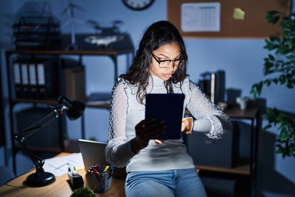 Young brazilian woman using touchpad at night working at the office checking the time on wrist watch, relaxed and confident 