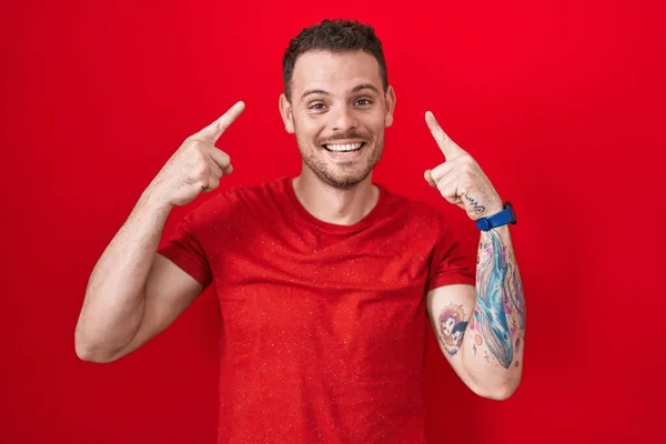 Kırmızı Arka Planda Duran Spanyol Genç Adam Gülümseyerek Iki Eliyle — Stok fotoğraf