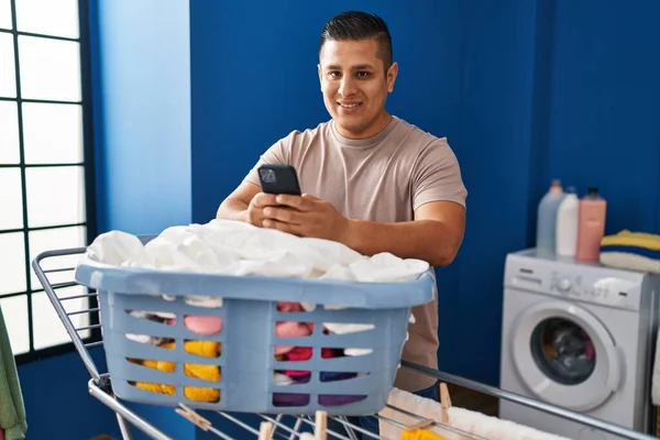 在洗衣房用智能手机把衣服挂在晾衣绳上的年轻拉丁男子 — 图库照片