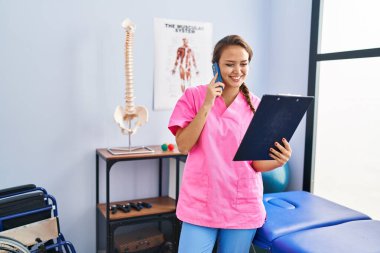 Young beautiful hispanic woman physiotherapist reading document talking on smartphone at rehab clinic